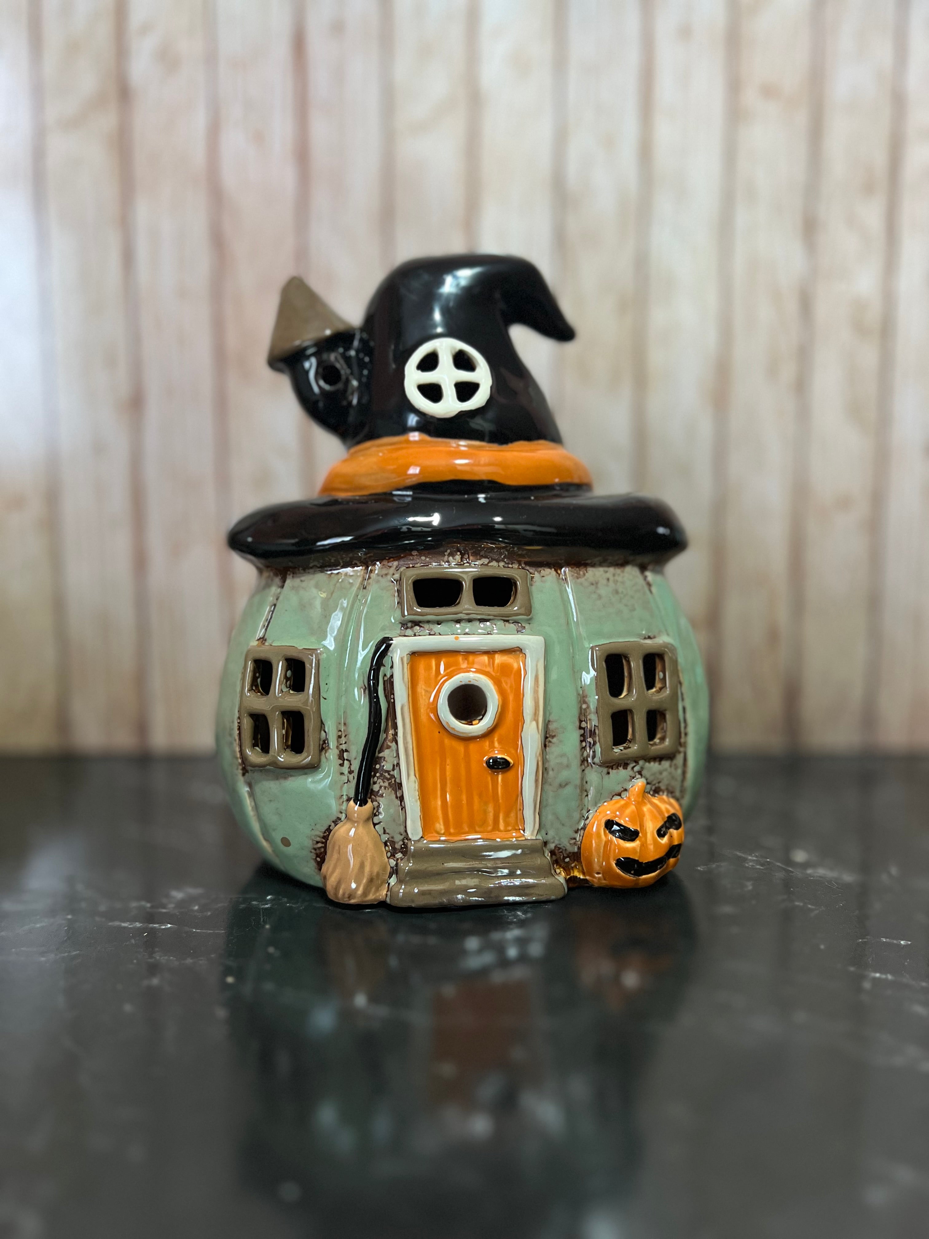 A small ceramic witch house figurine with a mint green body, orange door, black hat, and orange pumpkin accent, displayed on a dark reflective surface against a light wood background.