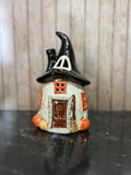 A small ceramic witch house figurine with a pointed black hat, orange window accents, a brown door, and pumpkin details, displayed on a dark reflective surface against a light wood background.