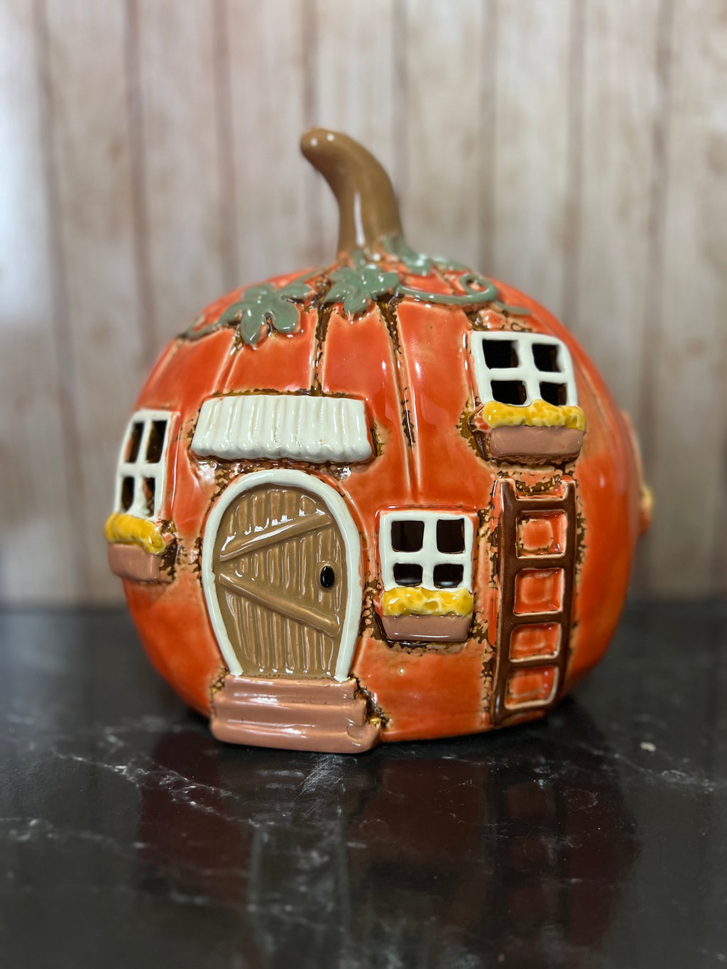 An orange ceramic pumpkin-shaped house with a brown stem, featuring a detailed brown door, white-framed windows, yellow flower boxes, and a small ladder, displayed on a dark reflective surface.