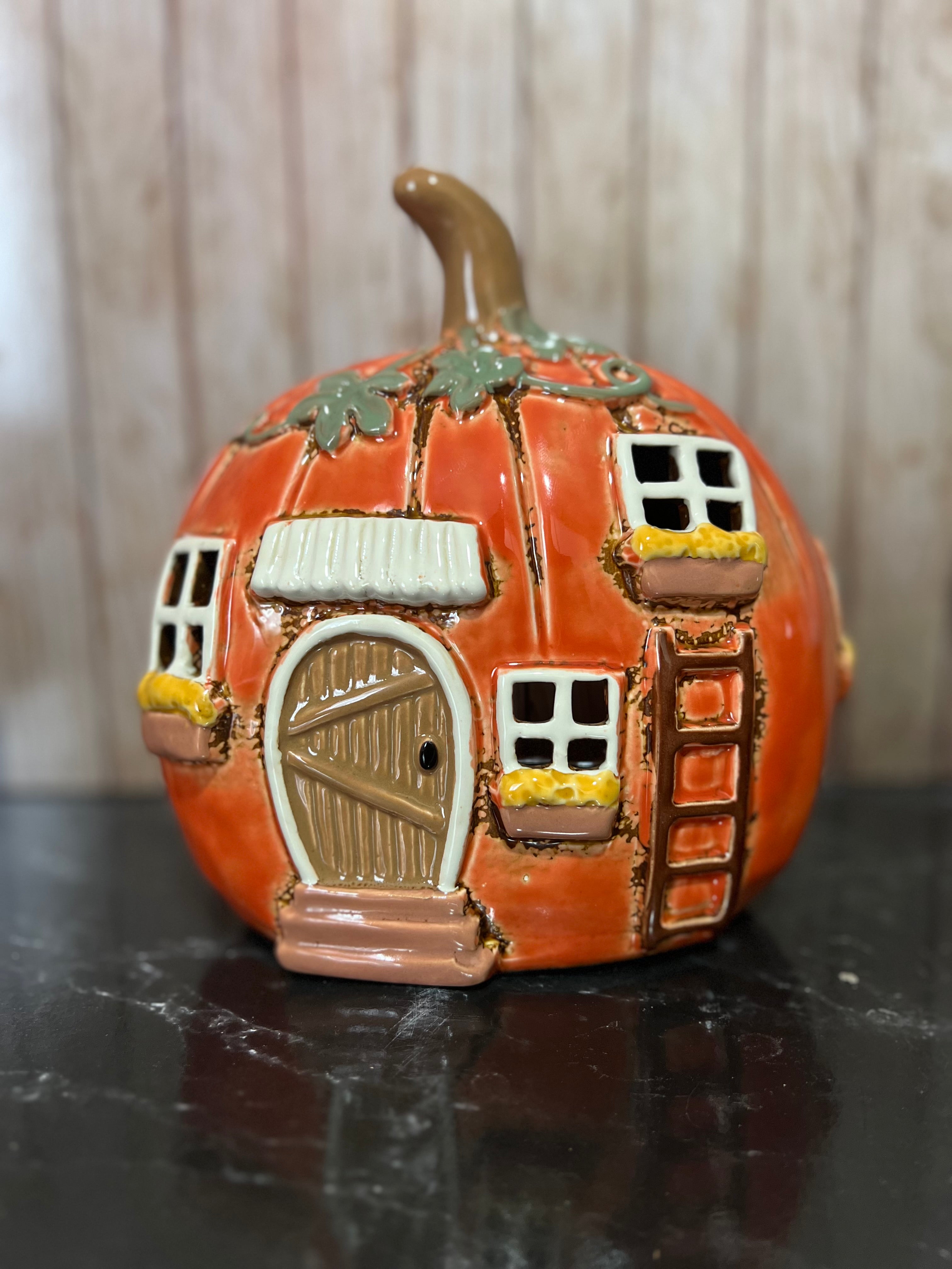 An orange ceramic pumpkin-shaped house with a brown stem, featuring a detailed brown door, white-framed windows, yellow flower boxes, and a small ladder, displayed on a dark reflective surface.