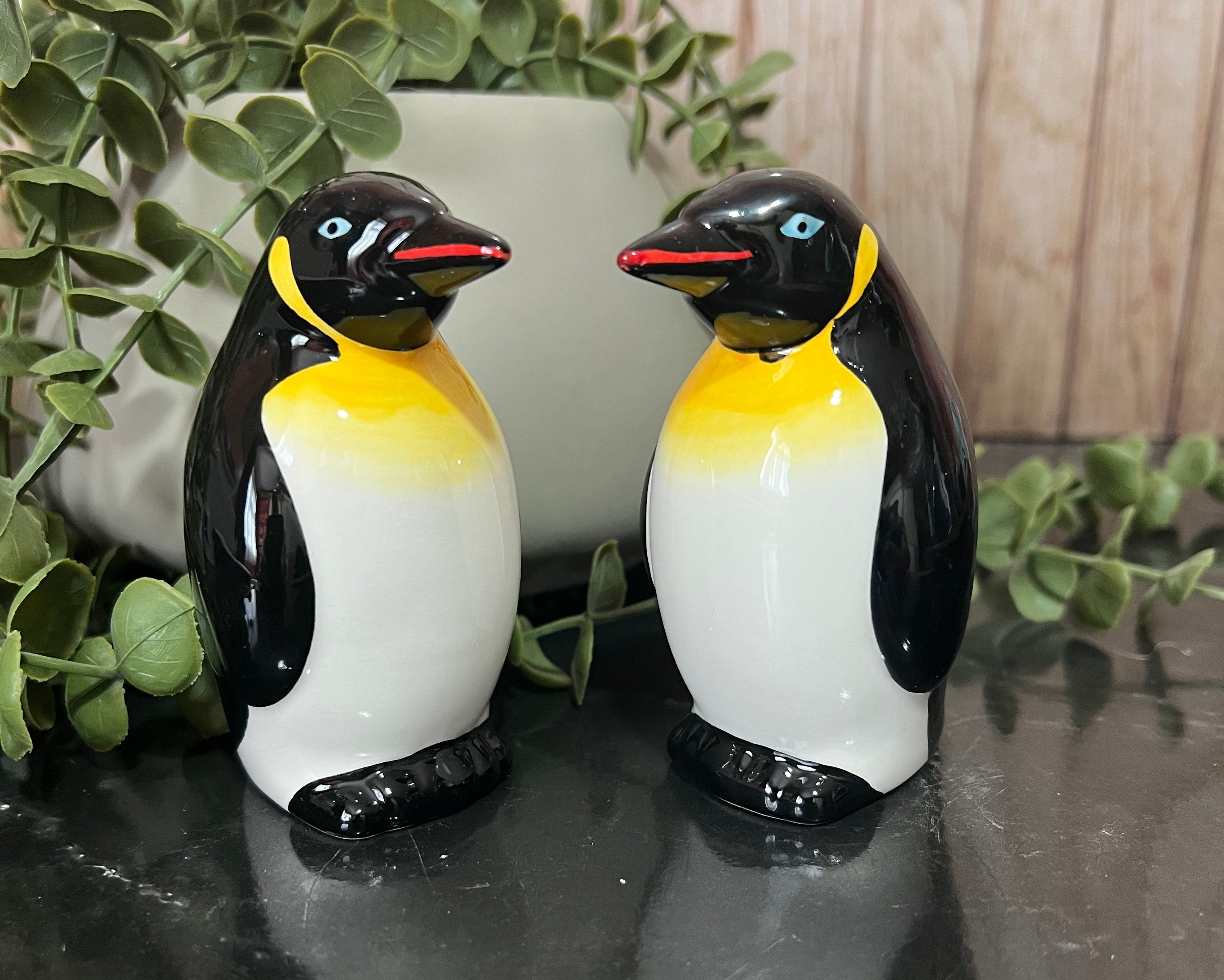 Two ceramic penguin figurines with black, white, and yellow coloring, standing on a dark reflective surface with green foliage and a light wood background.