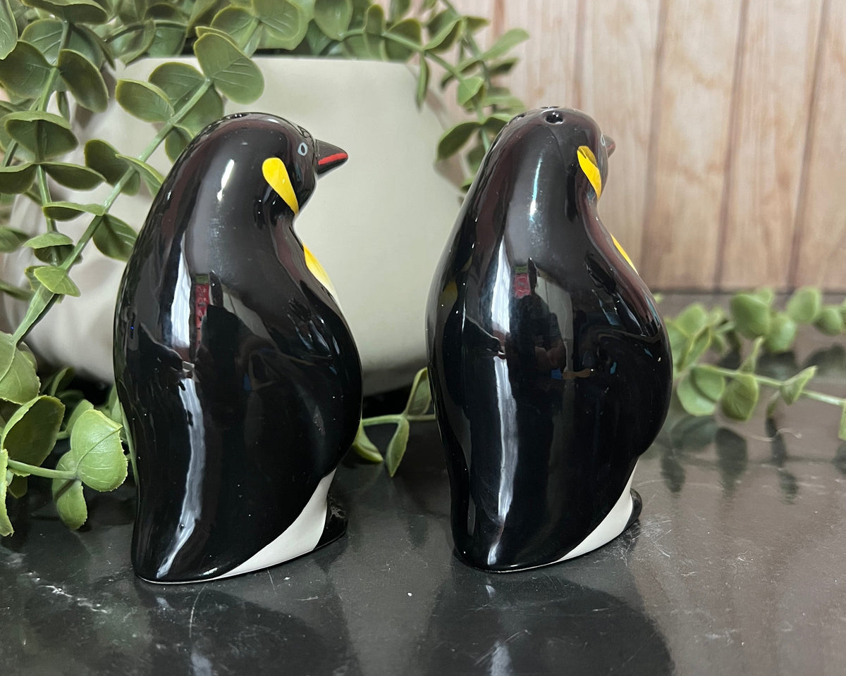 A pair of glossy black penguin figurines with yellow and white accents, displayed on a dark reflective surface with green foliage and a wooden background.
