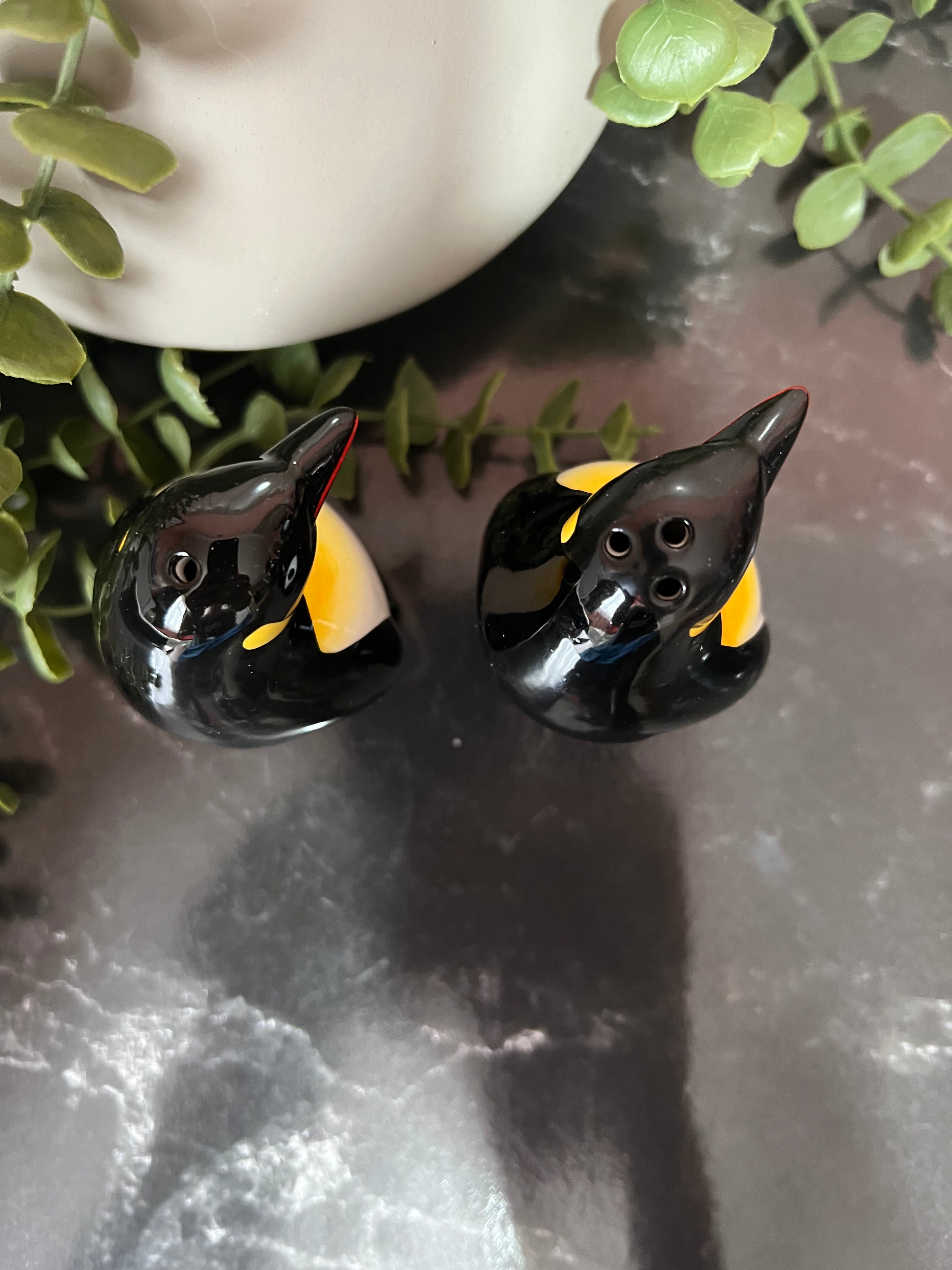 Two glossy black bird-shaped glass wind instruments with yellow and white accents, displayed on a dark marble surface with green leaves and a white ceramic vase in the background.