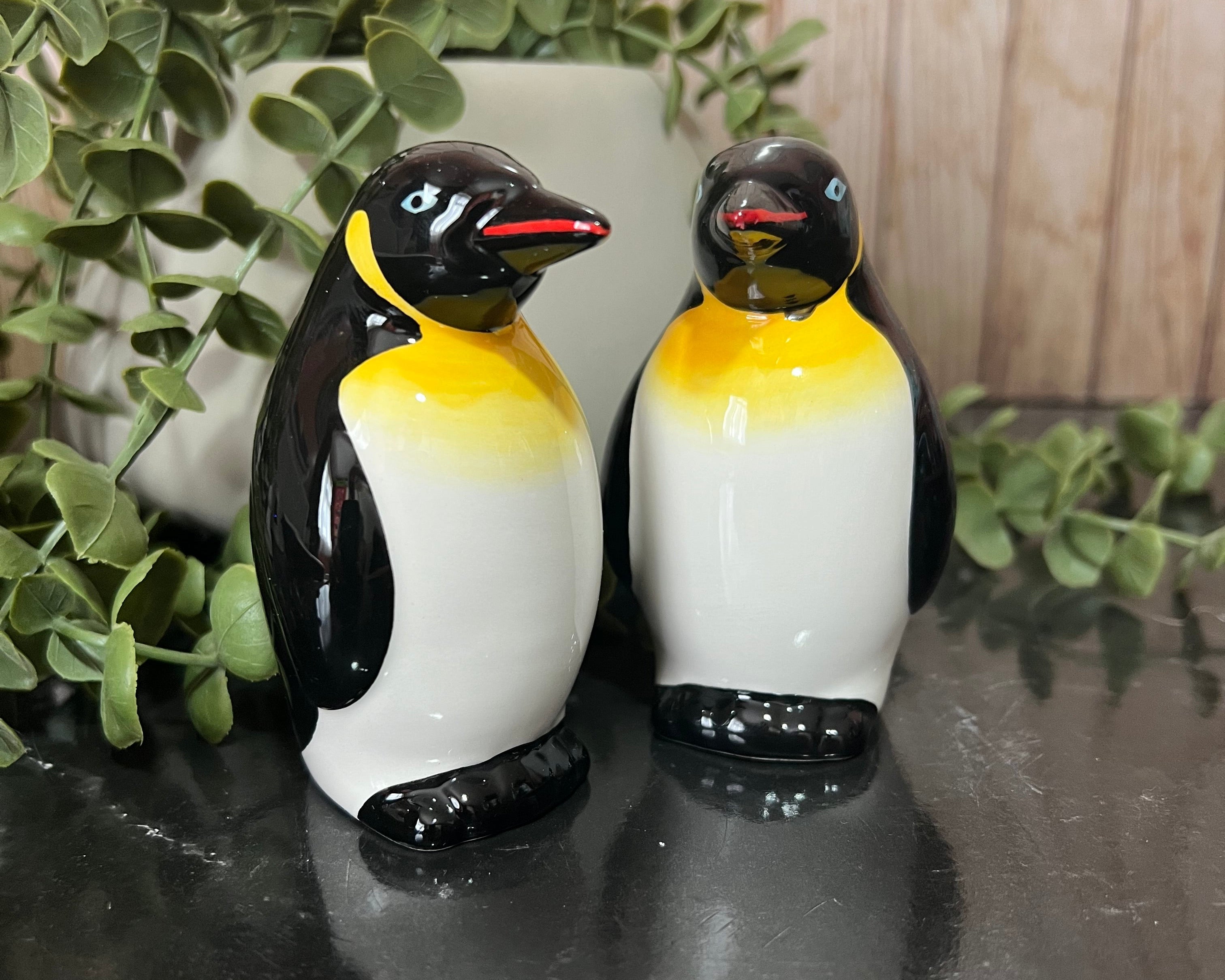 Two glossy penguin figurines with black, white, and yellow coloring, standing on a dark reflective surface with green foliage and a light wood background.